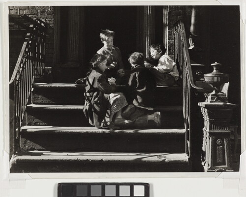 A black-and-white photograph of four children playing together on concrete steps with iron railings that cast striped shadows across them.