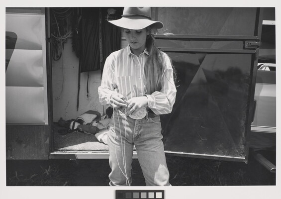A black-and-white photograph of a young White woman with long hair wearing western clothing cutting rope with a knife.