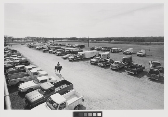 A black-and-white photograph of a single rider on horseback amid rows of parked cars and trucks.