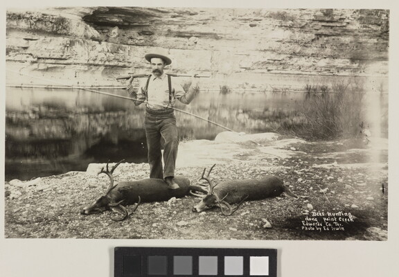 A black-and-white photograph of a White man racking a rifle across his shoulders while standing over two buck carcasses next to a creek.