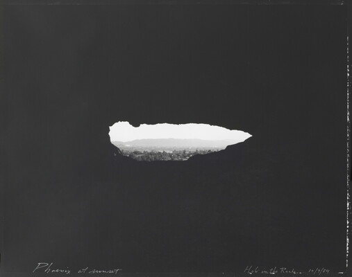 A black-and-white photograph of a hazy city beyond trees, mountains in the distance, viewed through a dark, irregular oblong shape.