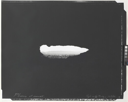 A black-and-white photograph of a hazy city beyond trees, mountains in the distance, viewed through a dark, irregular oblong shape.