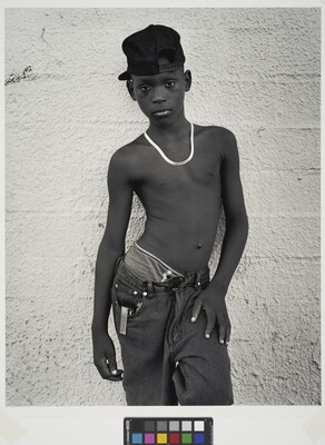 A black-and-white portrait photograph of a Black teenage boy wearing a backward baseball cap, a necklace, no shirt, and loose fitting jeans against a concrete wall.