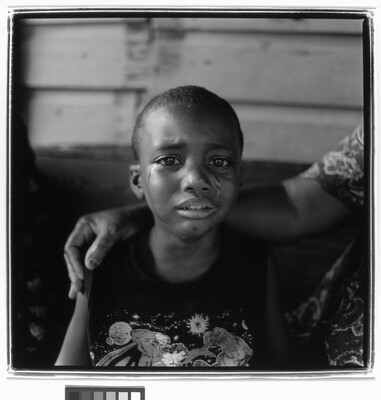 A black-and-white photograph of a young Black child with tears streaming down their face and someone's arm around their shoulders.