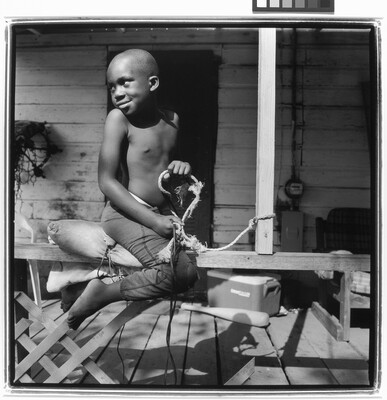 A black-and-white photograph of a young Black boy straddling the railing of a porch pretending to ride a horse.