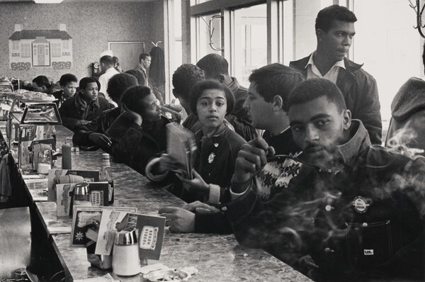 A black-and-white photograph of mostly young Black people and one White person at a crowded diner counter, talking and smoking.
