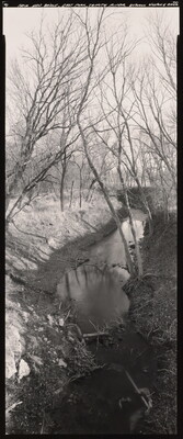 A vertically-oriented black-and-white photograph of a creek with bare trees on both sides.