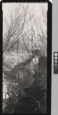 A vertically-oriented black-and-white photograph of a creek with bare trees on both sides.