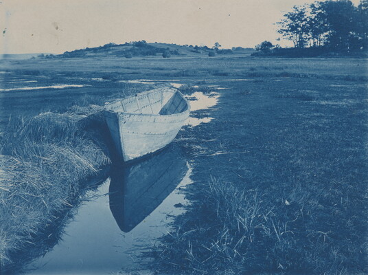 A blue-tinted photograph of an empty rowboat in a shallow creek surrounded by fields.