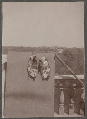 A black-and-white photograph of a pair of beaded moccasins hanging on a board that is leaning on a balustrade; tree-covered hills in the distance.