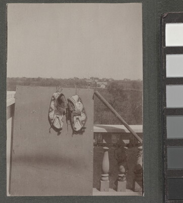 A black-and-white photograph of a pair of beaded moccasins hanging on a board that is leaning on a balustrade; tree-covered hills in the distance.