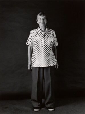 A black-and-white portrait photograph, in full length, of a smiling older White woman wearing  a short-sleeved polka dot shirt and long pants.