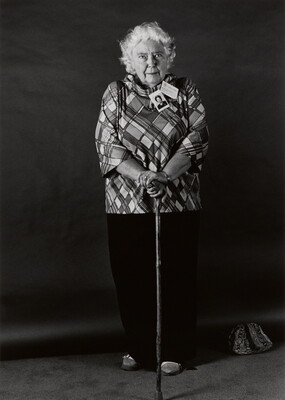A black-and-white portrait photograph, in full length, of a white-haired, elderly White woman wearing a plaid shirt, long dark pants, and resting her hands on a cane.