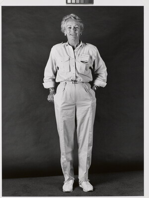 A black-and-white portrait photograph of a smiling middle-aged White woman with short hair wearing a button-down shirt with a name badge above the breast pocket, light colored pants, and sneakers.