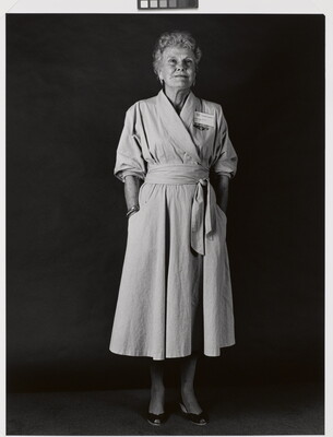 A black-and-white portrait photograph of an older White woman with short, light hair wearing a wrap dress with a name badge above her breast and her hands in her pockets.