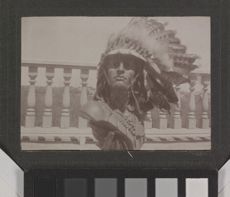 A sepia-toned photograph of a male Native American sculpture bust wearing a real feathered headdress and beads around the neck.