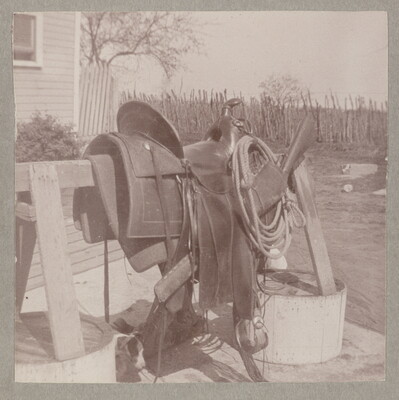 A sepia-toned photograph of the side view of a saddle and rope resting on a wooden sawhorse in a yard.