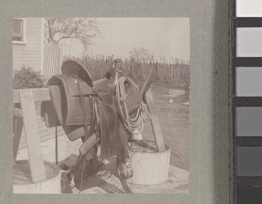 A sepia-toned photograph of the side view of a saddle and rope resting on a wooden sawhorse in a yard.