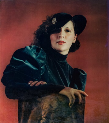 A color portrait photograph of a White woman with curly hair topped with a hat that covers one eye, wearing a green velvet dress with puffy sleeves, one arm resting on the back of a chair.
