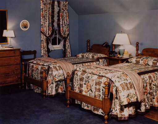 A color photograph of a bedroom with two twin beds, both with wood frames and matching floral print bedspreads that match the window curtains.