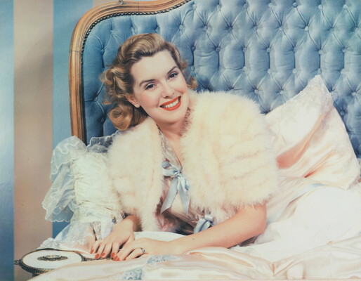 A color photograph of a smiling White woman wearing a white fur jacket, leaning on one elbow on a bed with a quilted blue headboard.