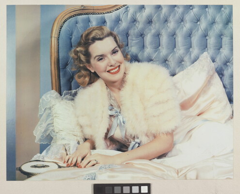 A color photograph of a smiling White woman wearing a white fur jacket, leaning on one elbow on a bed with a quilted blue headboard.