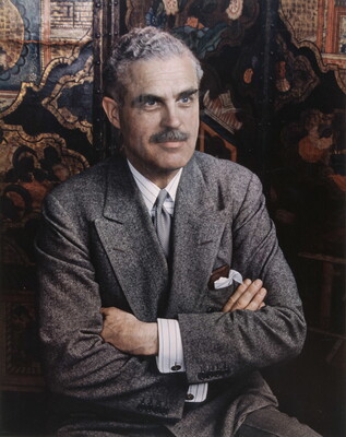 A color photograph of a seated White man with wavy gray hair and a mustache, wearing a gray suit and tie, arms crossed in front his chest.
