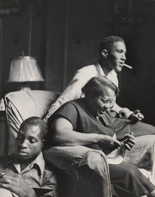 A black-and-white photograph of a Black woman knitting in an armchair, a young Black man smoking on the arm of her chair, and a young Black man seated on the floor reading.