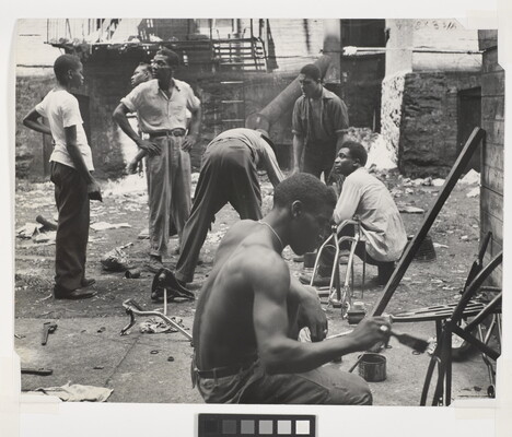 A black-and-white photograph of group of Black men talking and working in a rundown space littered with debris.