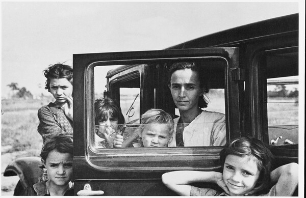 A black-and-white photograph of a White woman seated in the open door of a car with a young child on her lap; four other children stand behind and in front of the open car door.
