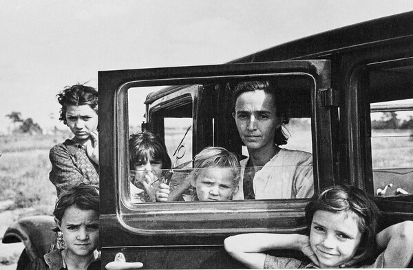 A black-and-white photograph of a White woman seated in the open door of a car with a young child on her lap; four other children stand behind and in front of the open car door.