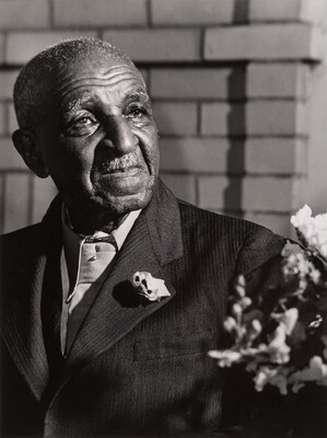 A black-and-white portrait photograph of an elderly Black man wearing a suit with a corsage, a brick wall in the background.