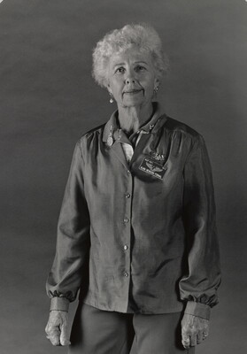 A black-and-white portrait photograph of an older White woman with short, curly gray hair wearing a long-sleeved button-down blouse.