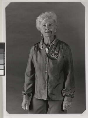 A black-and-white portrait photograph of an older White woman with short, curly gray hair wearing a long-sleeved button-down blouse.