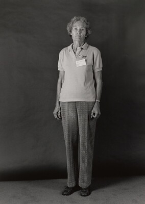 A black-and-white portrait photograph of an older White woman wearing a short sleeved polo shirt with a nametag pinned to her breast, and plaid trousers.