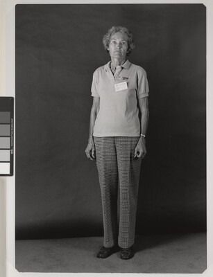 A black-and-white portrait photograph of an older White woman wearing a short sleeved polo shirt with a nametag pinned to her breast, and plaid trousers.