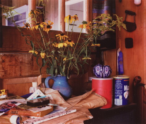 A color photograph of wilted yellow wildflowers in a blue ceramic pitcher on a cluttered table in a room with wood panel walls.