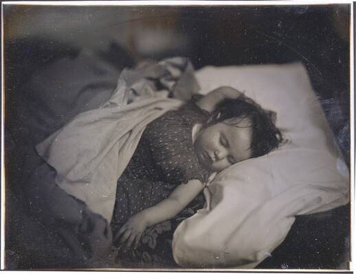 A black-and-white photograph of a White baby sleeping on their back, dark hair sticking to their head, in a bed with one hand above their head and the other at their side.