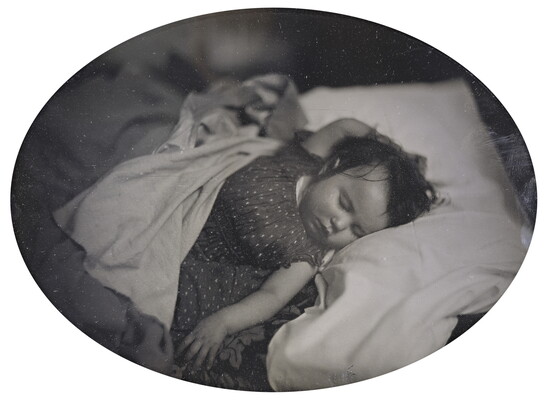A black-and-white photograph of a White baby sleeping on their back, dark hair sticking to their head, in a bed with one hand above their head and the other at their side.