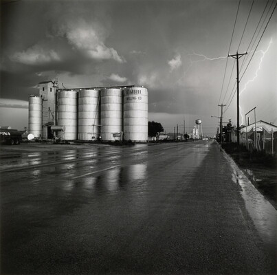 A black-and-white photograph of four grain elevators to the left of a wet road, a water tower in the far distance, and a flash of lighting behind power lines on the right.