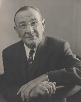 A black-and-white portrait photograph of an older, balding White man wearing glasses, a suit and tie, arms crossed over the back of a chair.