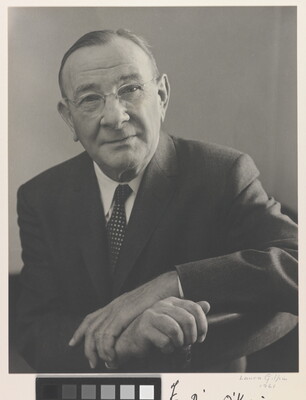 A black-and-white portrait photograph of an older, balding White man wearing glasses, a suit and tie, arms crossed over the back of a chair.