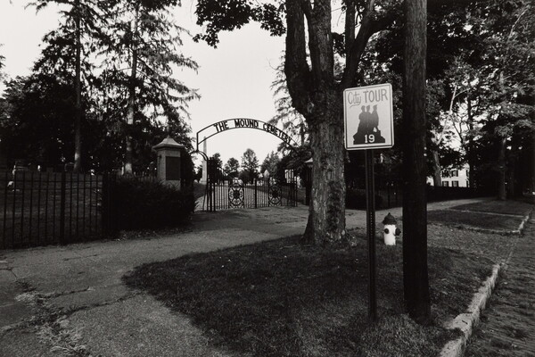 A black-and-white photograph taken from the street of a wrought iron gate at the entrance to a cemetery.