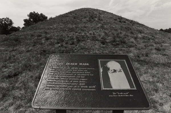 A black-and-white photograph of an outdoor informational plaque titled "The Death Mask" in front of a large grassy mound.