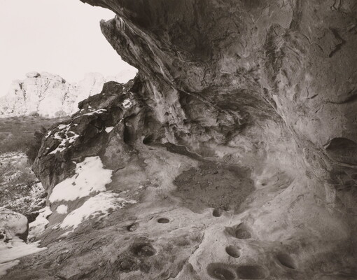A black-and-white photograph of an overhang in a rock wall; snow dapples the edge of the rocks.