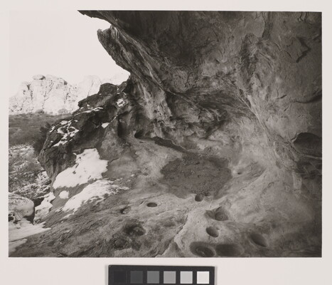 A black-and-white photograph of an overhang in a rock wall; snow dapples the edge of the rocks.