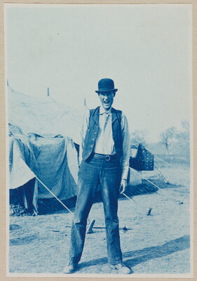 A black-and-white photograph of a White man with an animated expression in a bowler hat and vest standing in front of a tent.