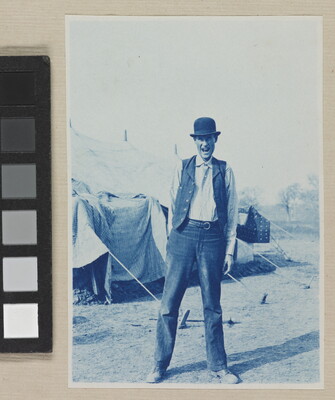 A black-and-white photograph of a White man with an animated expression in a bowler hat and vest standing in front of a tent.