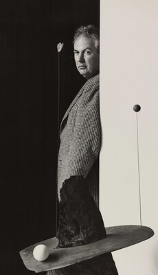A black-and-white photograph of a middle-aged White man in profile leaning against a wall; an abstract sculpture sits on a base in front of him.