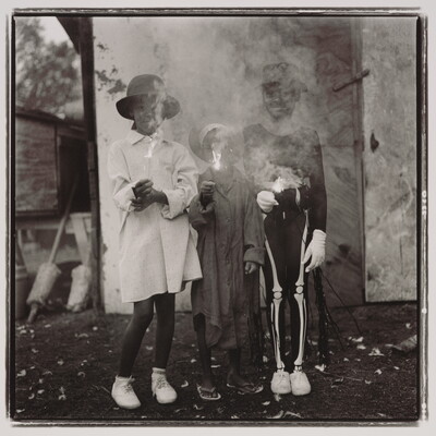 A black-and-white photograph of three Black children: one wearing a hat with a long white shirt, one a hat with a long dark shirt, and one with a hat wearing a skeleton costume; all are holding sparklers with the smoke obscuring their faces.